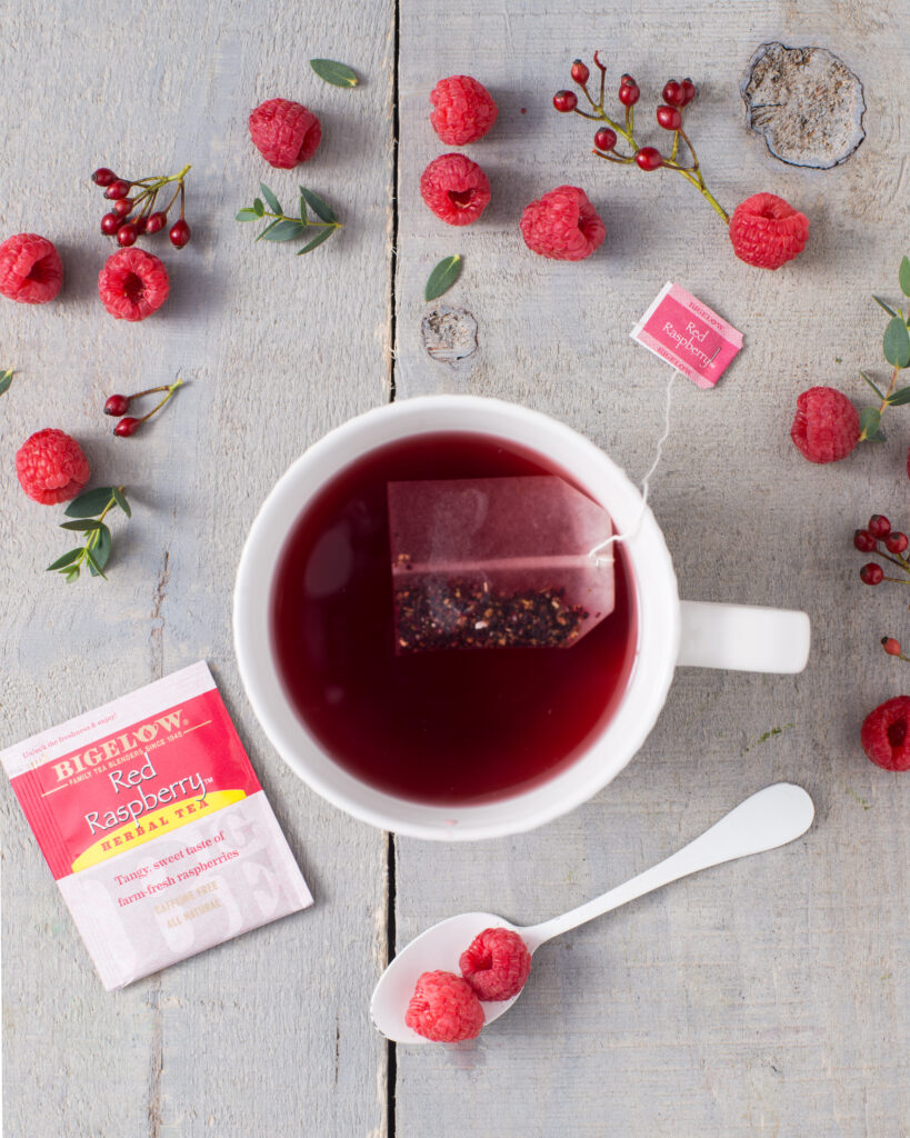 red raspberry tea bad next to white tea cup on table. raspberries arranged around cup.