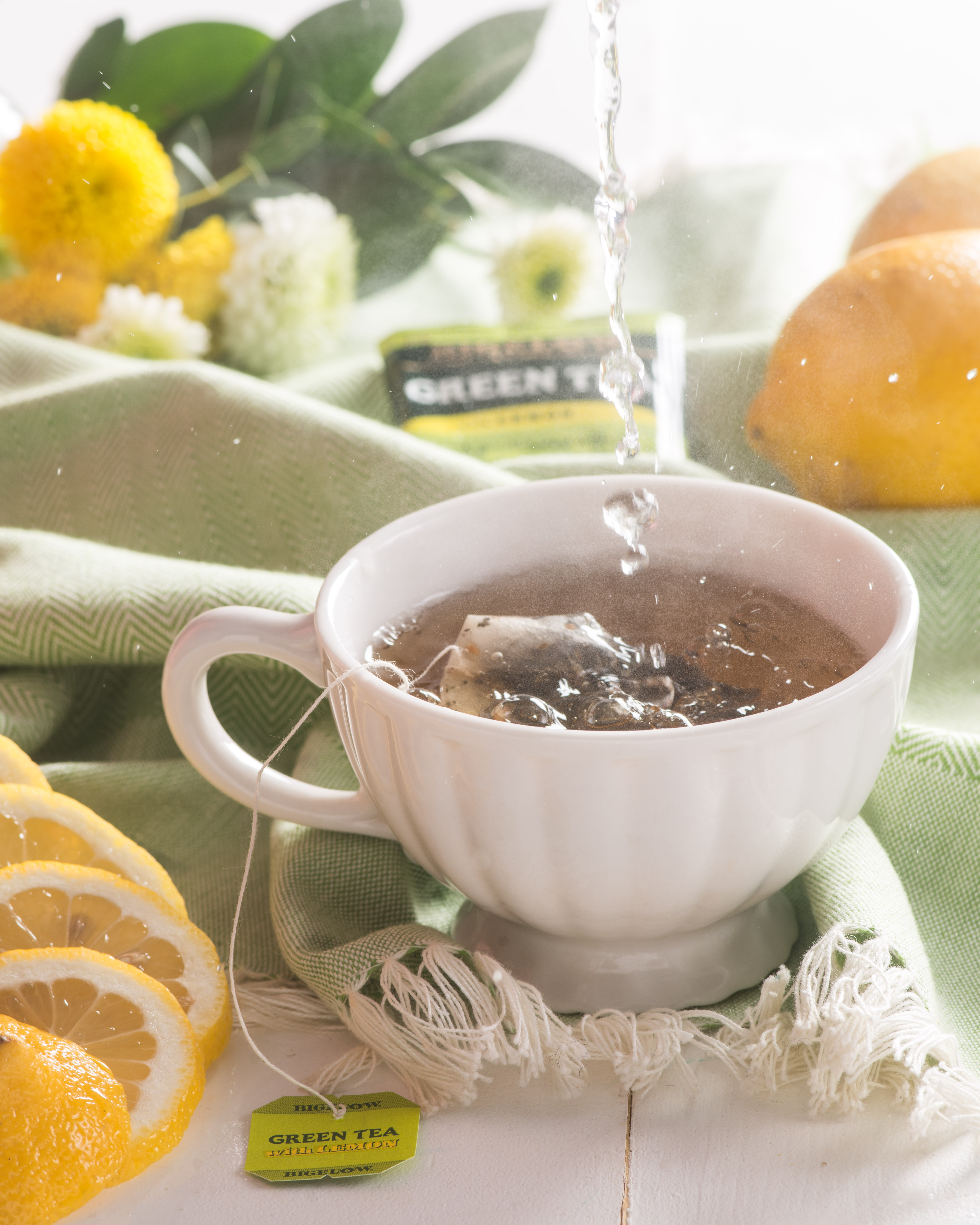 tea cup in foreground with lemons and green tea, tea bags in background with green table coth