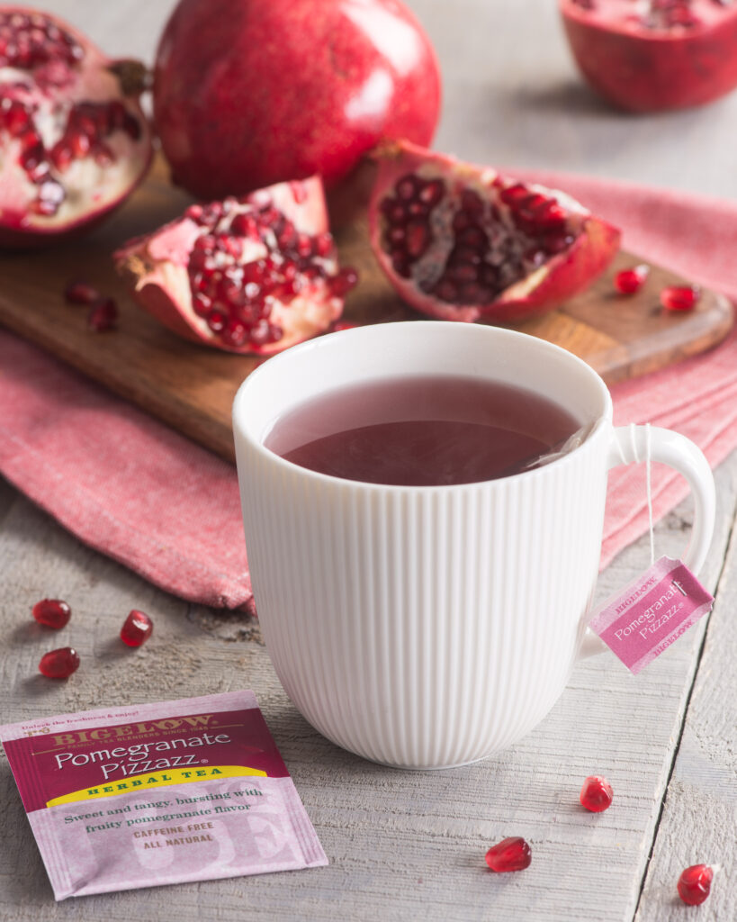 white mug of tea on table with pomegranate pizzaz pink and purple tea bag in foreground, and pomegranate on cutting board behind mug.