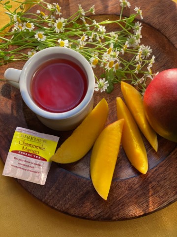 overhead view of mug filled with tea. sliced mangoes, tea bag, and chamomile flowers frame mug on table.